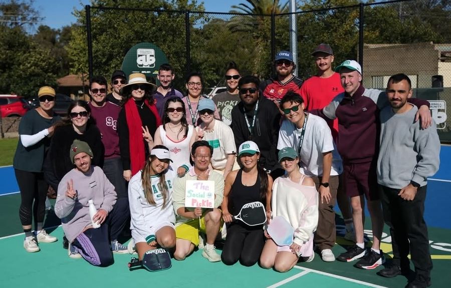 A large group of pickleball players gathered on court, one holding a "The Social Pickle" sign